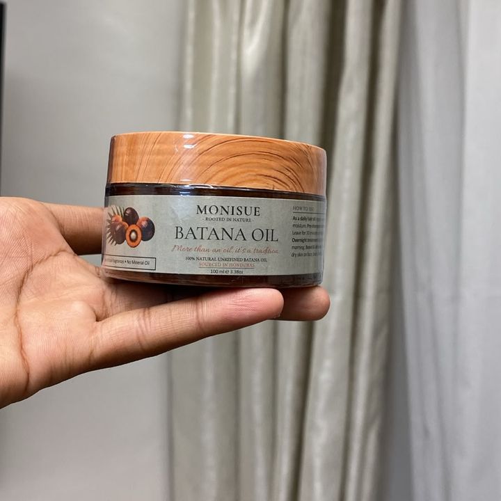 A hand holding a 100 ml jar of monisue batana oil with a wood-pattern lid. The label reads: 100% natural unrefined batana oil, sourced in Honduras.